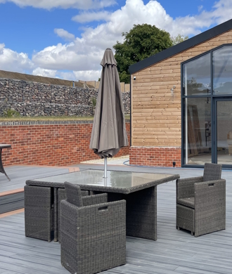 Showcasing Trex Composite Decking in Tiki Torch and Island Mist on a sunny day. Outdoor table and chair set situated on decking with clouds and blue sky to the top. 
